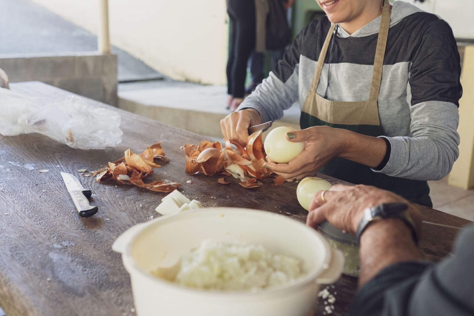 Two people peel and chop onions at a worn wooden table; one wears an apron.