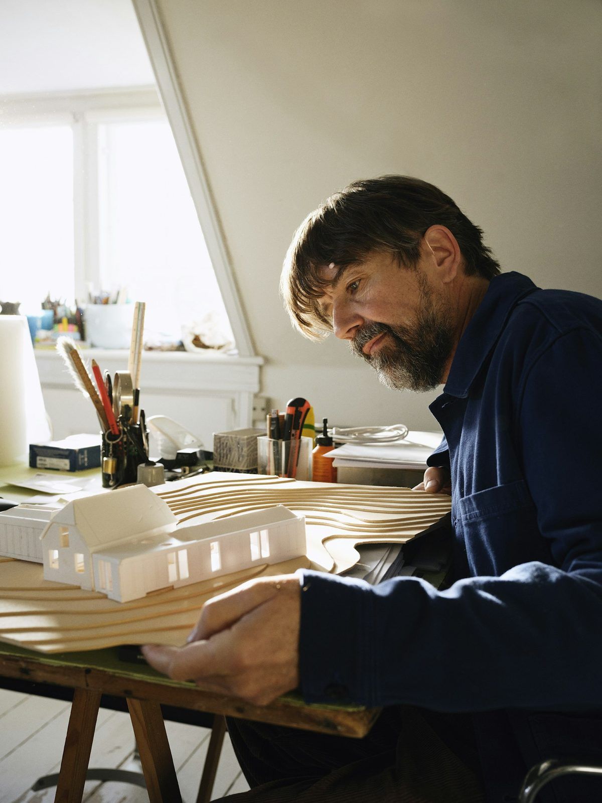 A man with a grey beard studies a white card model of a house on a contoured site board, by a skylight.